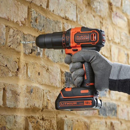 Photo of BLACK+DECKER BCD700S2AFC cordless drill in use drilling into a brick wall, 18V lithium battery visible.