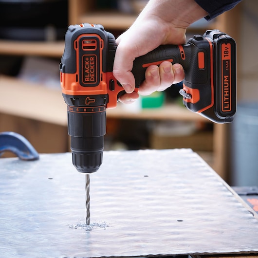 Photo of BLACKDECKER BDCHD18S1A cordless drill in use, drilling into metal sheet, black and orange design.