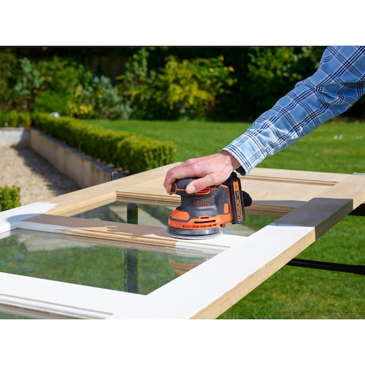 BLACKDECKER BDCROS18N cordless orange random orbital sander used on wooden window frame outdoors.
