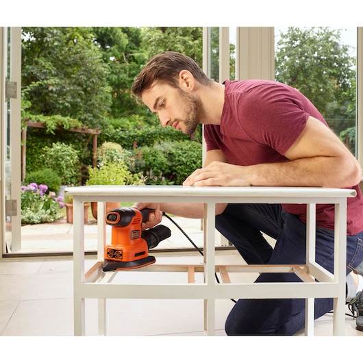 BLACKDECKER BEW200 sander in use sanding a white wooden table indoors near open doors.