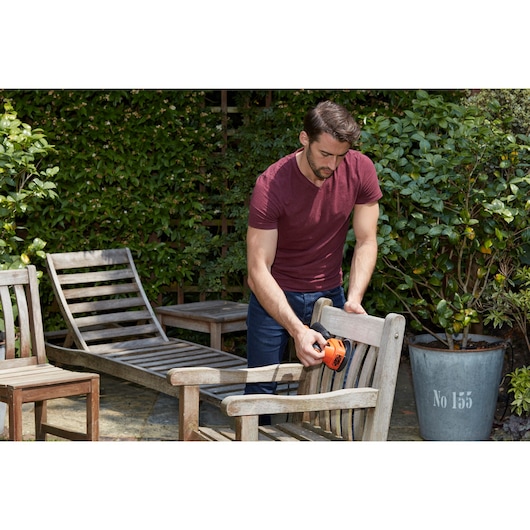 Person uses BLACKDECKER BEW200 detail sander on wooden outdoor chair in garden with other patio furniture visible.