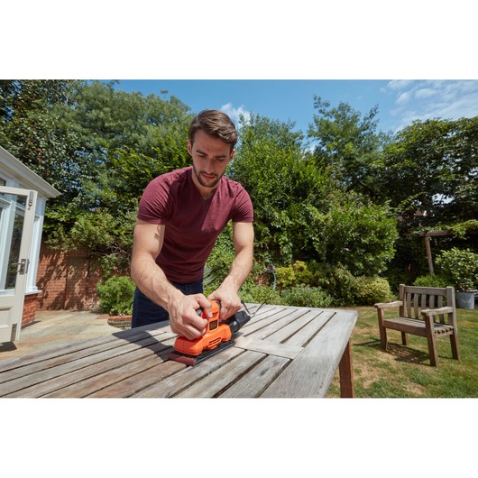 Photo of BLACK+DECKER BEW220 sander in use on a wooden table outdoors, person holding the tool.
