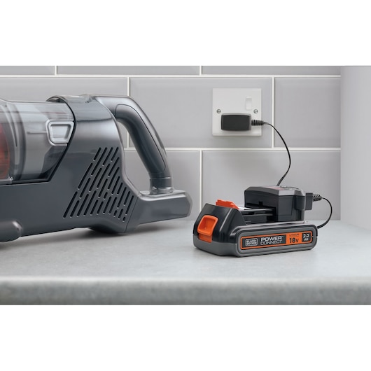 A BLACK+DECKER 18V lithium battery charger is placed on a countertop next to a cordless vacuum cleaner. The charger is plugged into a wall socket and is charging a battery. The scene is set against a tiled kitchen wall.