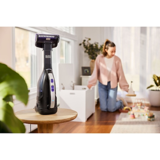 BLACK+DECKER hand vacuum DVD320BP on a wooden table in a bright room, play area visible in background.