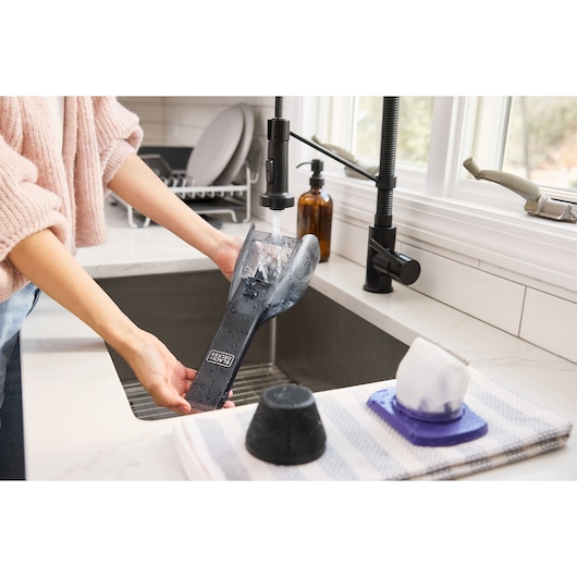 Person rinsing BLACK+DECKER DVD320BP vacuum filter under kitchen faucet, filter parts drying on towel nearby.