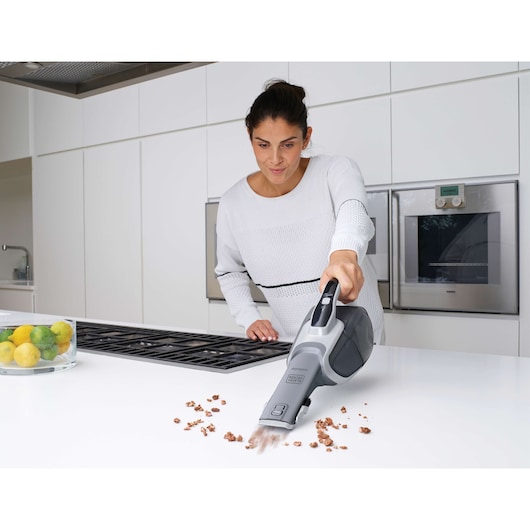 A person using a BLACK+DECKER handheld vacuum cleaner (DVJ215J) to clean crumbs from a white kitchen countertop. The kitchen is modern with built-in appliances and a bowl of lemons and limes is visible on the counter.