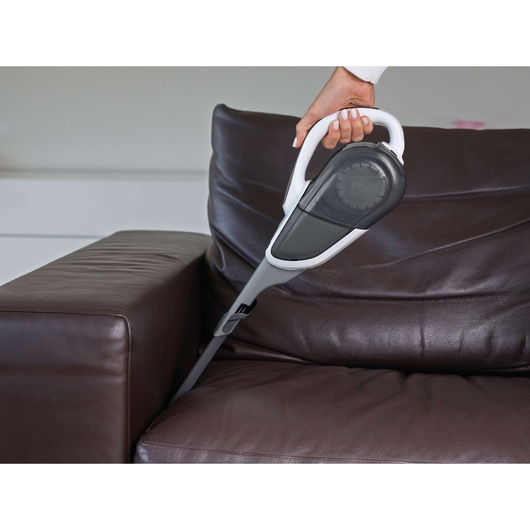 A hand holding a BLACK+DECKER handheld vacuum cleaner, cleaning the crevice between the seat and armrest of a dark brown leather sofa.