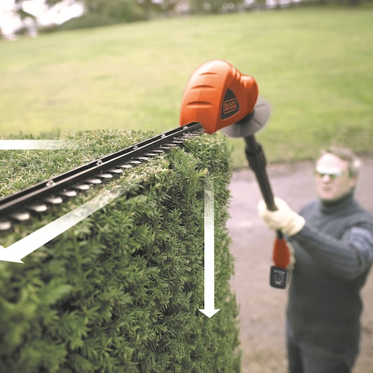 BLACKDECKER GTC1843L20 hedge trimmer in use cutting tall green hedge outdoors.