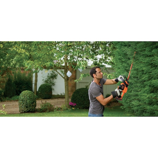 Photo of person using BLACK+DECKER GTC18452PCB cordless hedge trimmer to cut green hedge in garden.