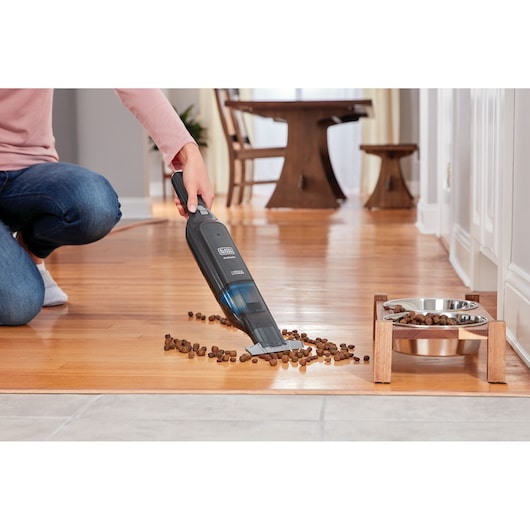 BLACKDECKER HLVC320J11 handheld vacuum cleaning pet food from hardwood floor near pet bowls.