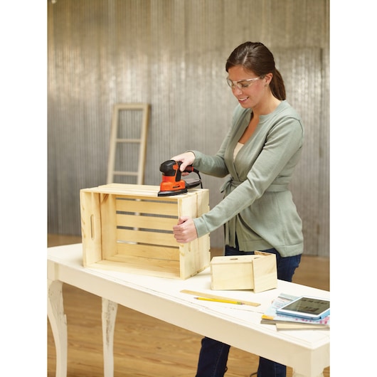Photo of person sanding a wooden crate with BLACKDECKER KA2500K sander on a table in a workshop.