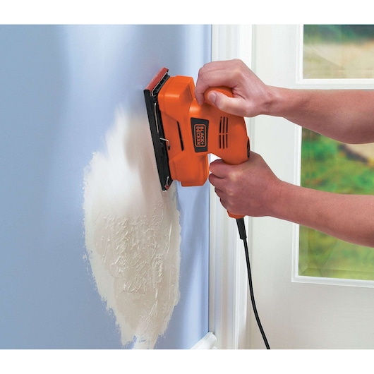 A person is using an orange BLACK+DECKER electric sander to smooth a patch of plaster on a blue wall near a white door.