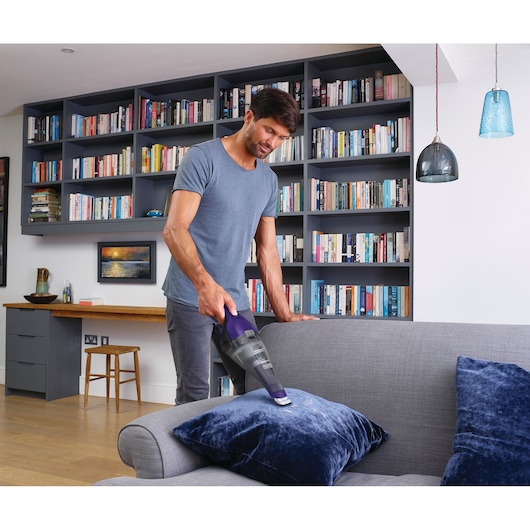 Person using BLACK+DECKER NVC215WP handheld vacuum to clean a blue cushion on a gray sofa in a modern living room.