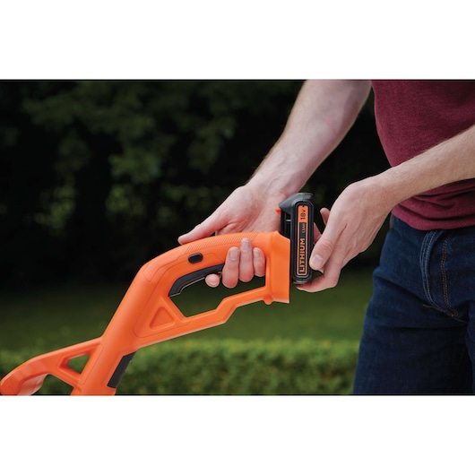 Photo of BLACKDECKER ST1823 orange cordless grass trimmer handle with battery being inserted, outdoor setting.