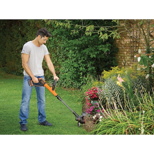 BLACKDECKER STC1820PCB grass trimmer used in garden next to flowerbed, orange and black cordless tool.